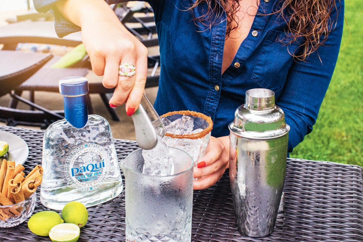 Woman pouring PaQui Tequila cocktail from shaker into glass, outdoor setting